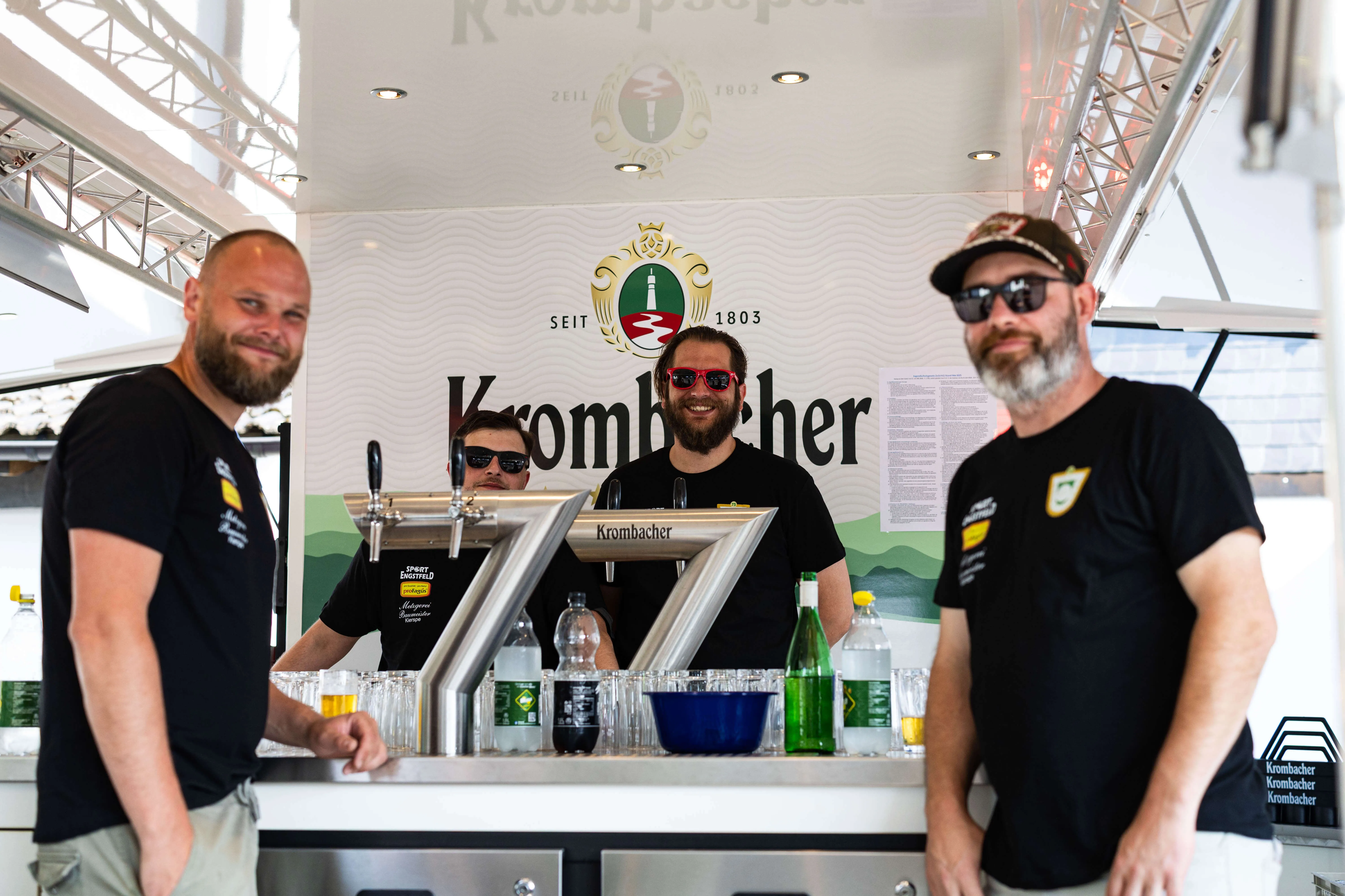 Drei Männer bedienen einen Krombacher-Bierstand auf dem Schützenplatz während eines Jubiläumsfestes.