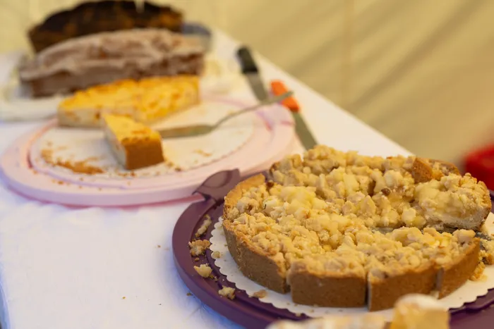 Verschiedene Kuchen, darunter ein Streuselkuchen auf einem lila Teller und ein gelber Kuchen, stehen auf einem weiß gedeckten Tisch.