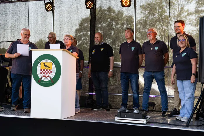 Gruppe von acht Personen steht auf einer Bühne vor einem Podest mit dem Vereinslogo des KKSV Meinerzhagen und singt gemeinsam.