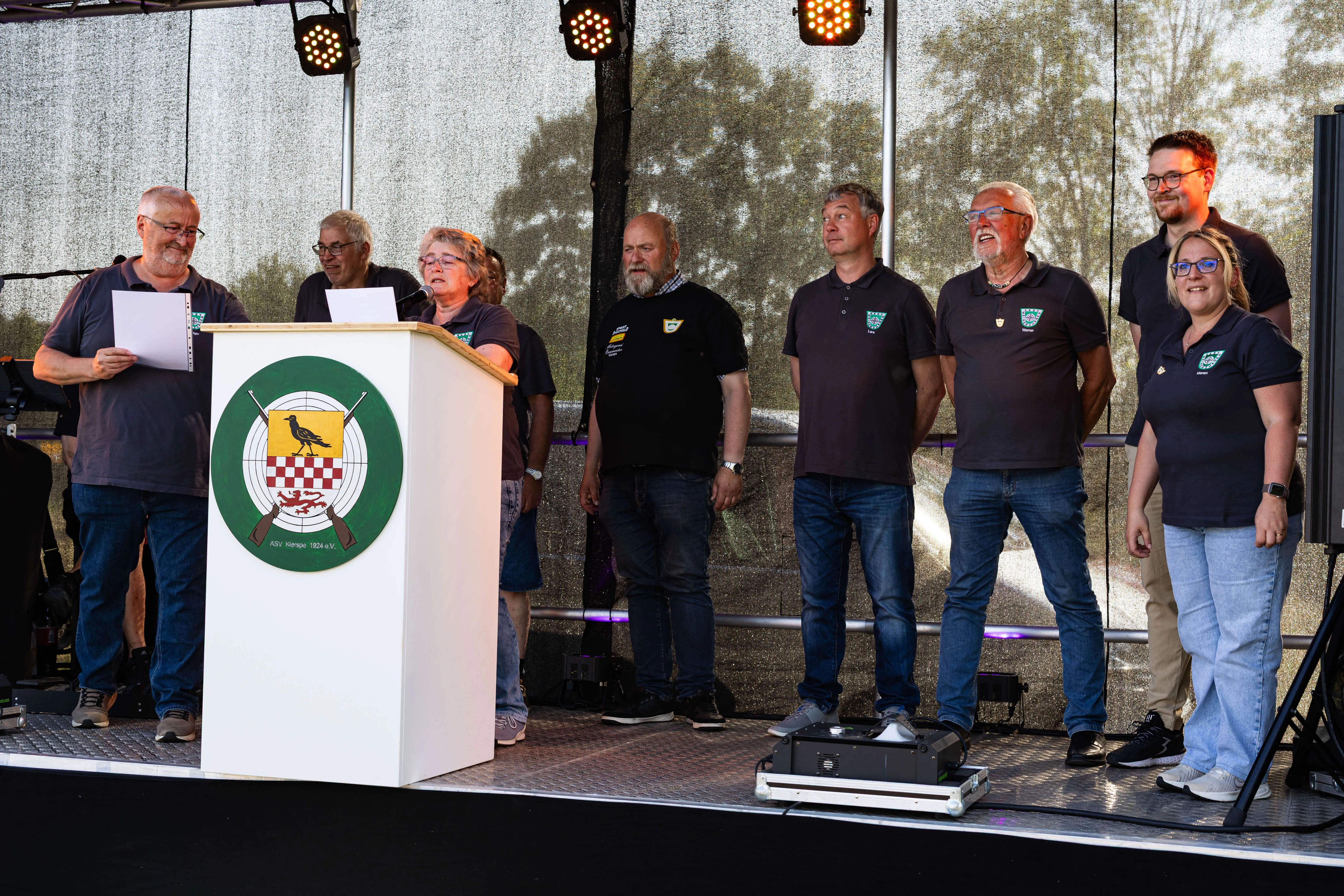 Gruppe von acht Personen steht auf einer Bühne vor einem Podest mit dem Vereinslogo des KKSV Meinerzhagen und singt gemeinsam.