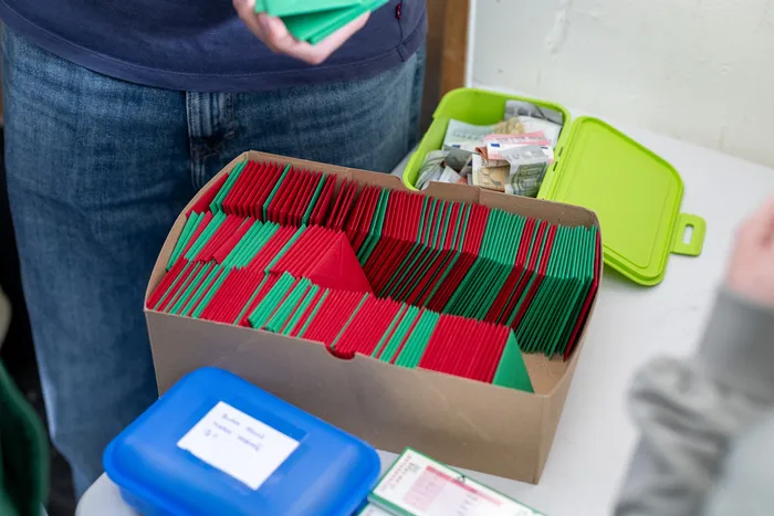 Gestapelte Sammelkarten in Rot und Grün in einem Karton, neben einer grünen Kasse mit Geldscheinen. Im Vordergrund eine blaue Kasse und Zettel.