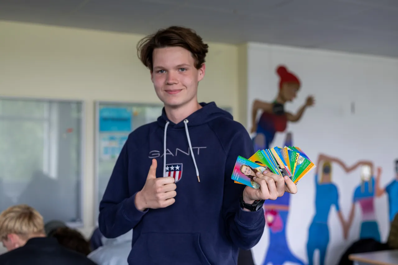 Ein junger Mann mit braunen Haaren und blauem Hoodie zeigt mit erhobenem Daumen eine Handvoll Sammelkarten mit Porträts von Lehrern. Im Hintergrund sind weitere Schüler und eine Wanddekoration mit einer stilisieren Figur zu sehen.