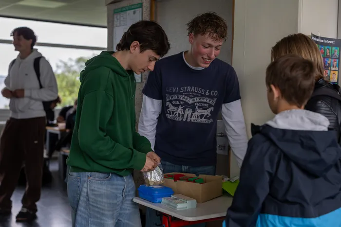 Junge Schüler verkaufen an einem Tisch Sammelkarten von Lehrern, um Geld für ihre Abikasse zu sammeln. Ein Schüler in grünem Hoodie und ein anderer mit einem Levi Strauss T-Shirt sind dabei zu sehen.