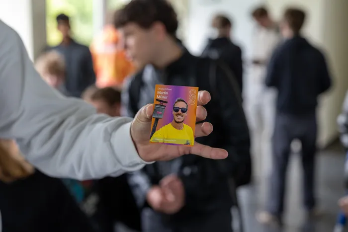 Eine Hand hält eine Sammelkarte mit dem Porträt von Martin Müller, einem Lehrer des Anne-Frank-Gymnasiums, vor einer Menschenmenge. Die Karte ist Teil einer Aktion zur Finanzierung der Abi-Kasse der Q1.