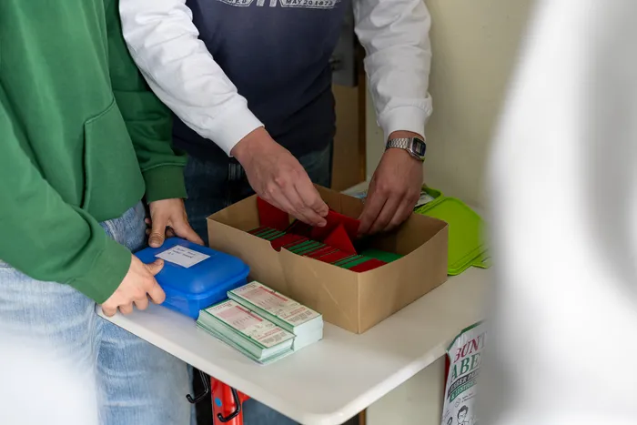 Zwei Personen sortieren rote und grüne Sammelkarten auf einem Tisch, um Spenden für den Abi-Kassen des Anne-Frank-Gymnasiums zu sammeln. Die Karten zeigen Lehrer des Gymnasiums.