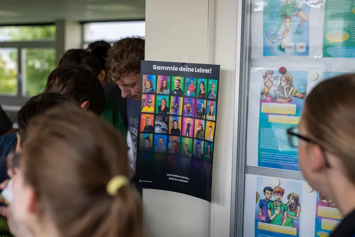 Ein Plakat bewirbt Lehrer-Sammelkarten, die Schüler des Anne-Frank-Gymnasiums für ihre Abi-Kasse verkaufen. Das Plakat zeigt Porträts verschiedener Lehrer.