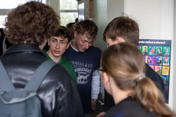 Eine Gruppe von Schülern steht um einen Tisch und betrachtet Sammelkarten mit Porträts von Lehrern. Im Hintergrund ist ein Plakat mit der Aufschrift „Sammele deine Lehrer!“ zu sehen.