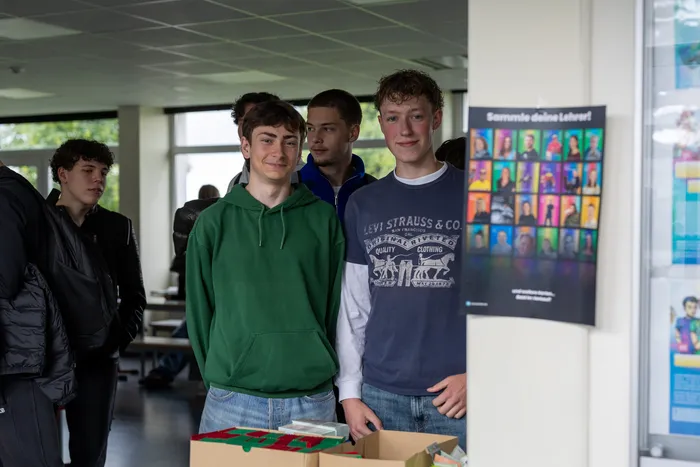 Zwei junge Männer stehen hinter einem Tisch mit gestapelten Kartenschachteln und verkaufen Sammelkarten mit Porträts von Lehrern. Im Hintergrund stehen weitere Schüler und ein Plakat mit den gleichen Lehrerporträts.