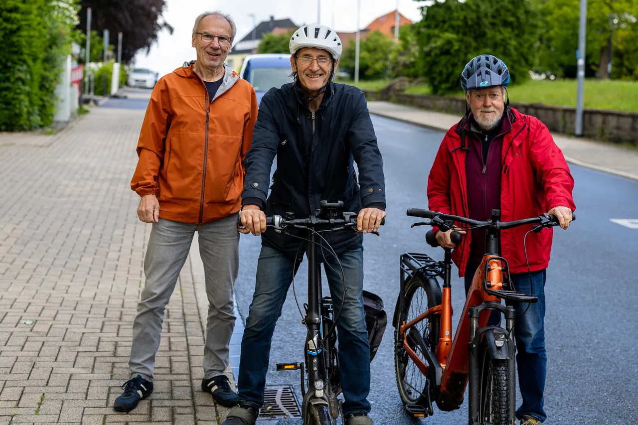 Drei ältere Männer stehen nebeneinander auf einem Bürgersteig mit Fahrrädern. Der Mann in der Mitte trägt einen Helm und sitzt auf einem Fahrrad, die anderen beiden stehen daneben.