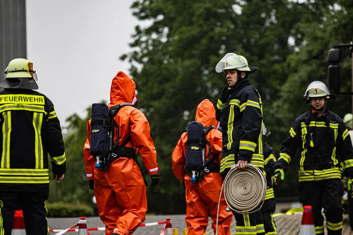 Feuerwehrleute in Schutzanzügen und Einsatzkleidung stehen auf einem Parkplatz vor einer grauen Gebäudefassade. Ein Feuerwehrmann trägt eine Schlauchrolle. Im Hintergrund sind weitere Einsatzkräfte und Absperrungen zu sehen.