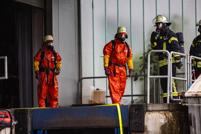 Drei Einsatzkräfte in orangefarbenen Schutzanzügen und mit Atemschutzmasken stehen auf einer erhöhten Plattform vor einem grauen Gebäude. Im Hintergrund sind weitere Feuerwehrleute in voller Montur zu sehen.