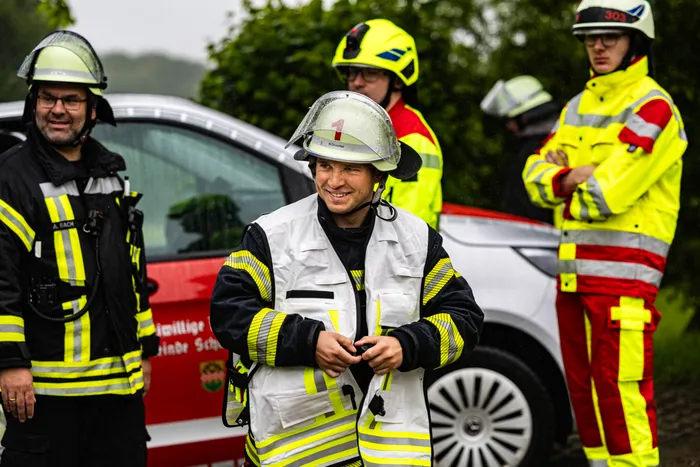 Ein Feuerwehrmann in voller Montur lächelt in die Kamera, während im Hintergrund weitere Einsatzkräfte und Einsatzfahrzeuge zu sehen sind. Der Einsatz findet im Gewerbepark Schalksmühle statt.
