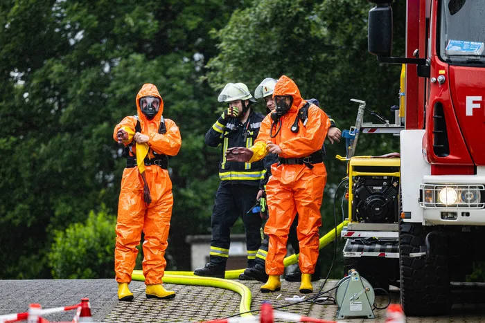 Einsatzkräfte in orangefarbenen Schutzanzügen mit Gasmasken stehen vor einem roten Feuerwehrauto. Im Hintergrund ist ein weiterer Feuerwehrmann in voller Montur zu sehen.