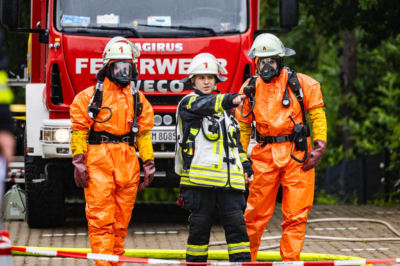 Drei Einsatzkräfte in orangefarbenen Schutzanzügen und Atemschutzmasken stehen vor einem roten Feuerwehrauto. Ein weiterer Feuerwehrmann zeigt in eine Richtung, während er eine Schutzausrüstung trägt. Im Hintergrund ist Absperrband zu sehen.