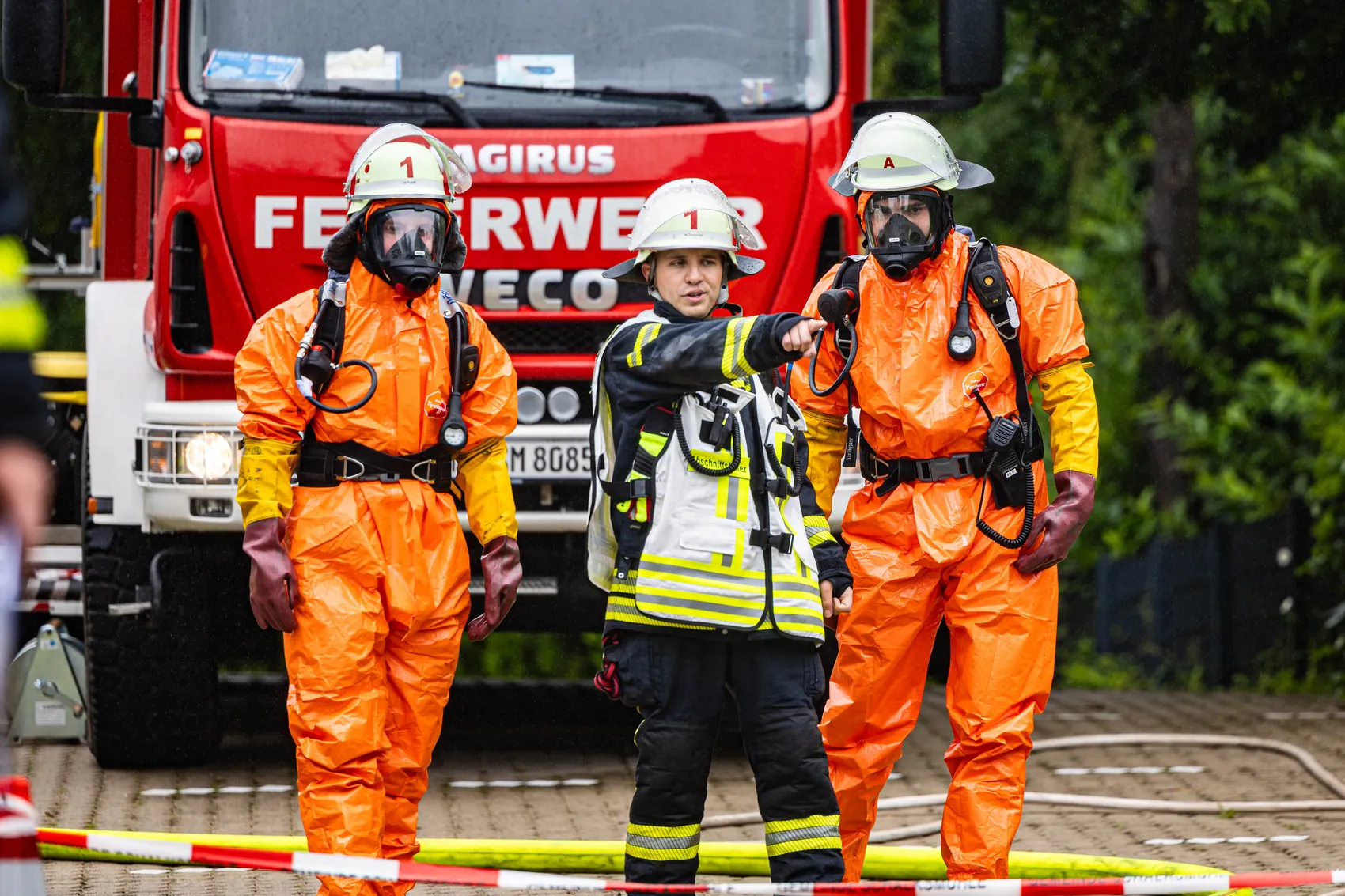 Drei Einsatzkräfte in orangefarbenen Schutzanzügen und Atemschutzmasken stehen vor einem roten Feuerwehrauto. Ein weiterer Feuerwehrmann zeigt in eine Richtung, während er eine Schutzausrüstung trägt. Im Hintergrund ist Absperrband zu sehen.