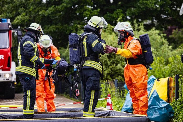 Feuerwehrleute in voller Schutzausrüstung besprechen während eines Gefahrguteinsatzes im Gewerbepark Schalksmühle die Vorgehensweise. Ein Teammitglied trägt eine orangefarbene Schutzanzug und zeigt auf eine Karte oder ein Diagramm.