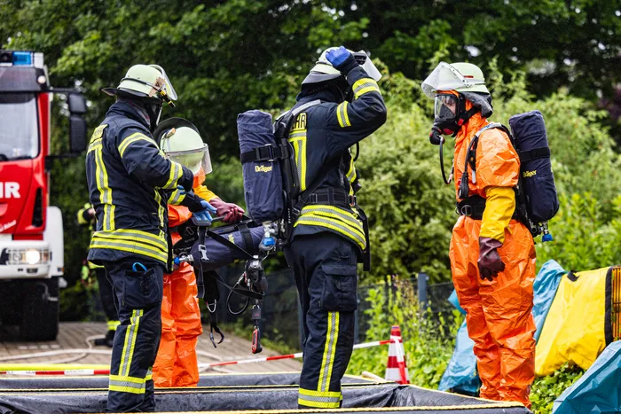 Vier Feuerwehrleute in voller Schutzausrüstung stehen im Kreis und bereiten sich auf einen Einsatz vor. Zwei tragen orangefarbene Schutzanzüge, die anderen dunkle. Im Hintergrund ist ein rotes Feuerwehrauto zu sehen.