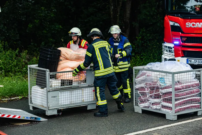 Feuerwehrleute tragen eine orangefarbene Puppe in einem Metallkorb zu einem roten Feuerwehrauto. Im Hintergrund sind weitere Einsatzkräfte und ein weiterer Metallkorb mit Trümmern zu sehen.
