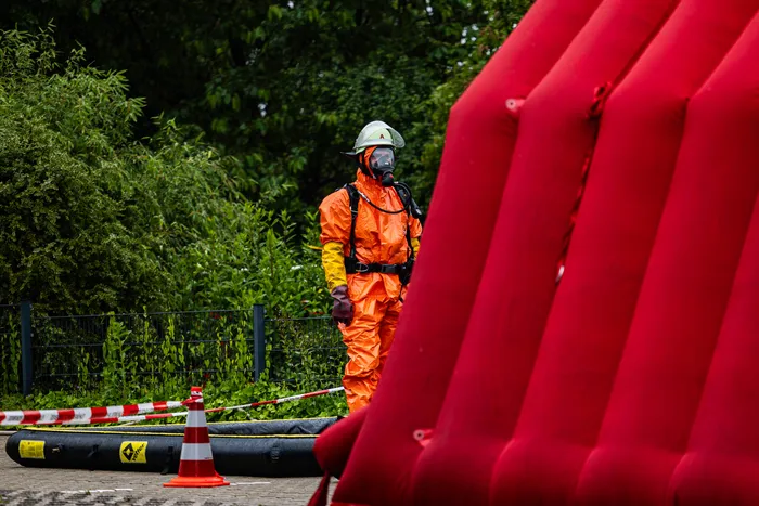 Ein Einsatzkraft in einem orangefarbenen Schutzanzug mit Helm und Atemschutzmaske steht neben einem roten aufblasbaren Zelt. Im Hintergrund ist eine Absperrung und Grün zu sehen.