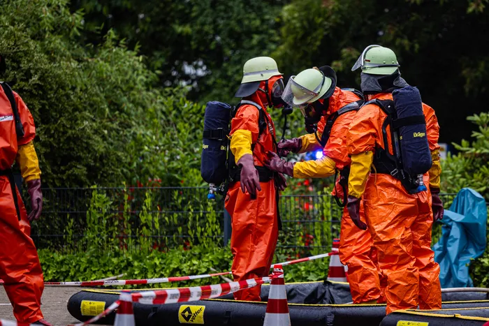 Einsatzkräfte in orangefarbenen Schutzanzügen mit Atemschutzmasken und Rückenbeatmung arbeiten während eines Gefahrguteinsatzes in Schalksmühle zusammen. Im Vordergrund befindet sich eine schwarze Absperrung mit Warnhinweisen.
