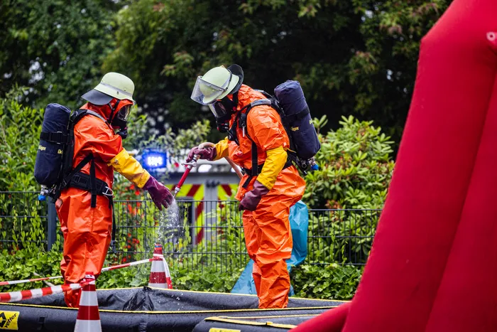 Zwei Einsatzkräfte in orangefarbenen Schutzanzügen und Atemschutzgeräten sind bei einem Gefahrguteinsatz im Gewerbepark Schalksmühle zu sehen. Sie stehen neben einer gelben Absperrbänderung und einer Wasserfontäne.