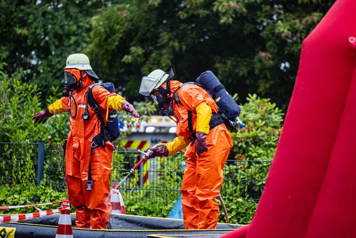 Zwei Einsatzkräfte in orangefarbenen Schutzanzügen mit Helmen und Atemschutzgeräten sind bei einem Gefahrguteinsatz im Gewerbepark Schalksmühle im Einsatz. Einer der Einsatzkräfte hält eine Sprühlanze.