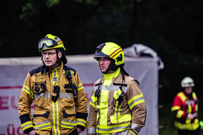 Zwei Feuerwehrleute in voller Schutzkleidung stehen während eines Gefahrguteinsatzes im Gewerbepark Schalksmühle nebeneinander. Im Hintergrund sind weitere Einsatzkräfte zu sehen.