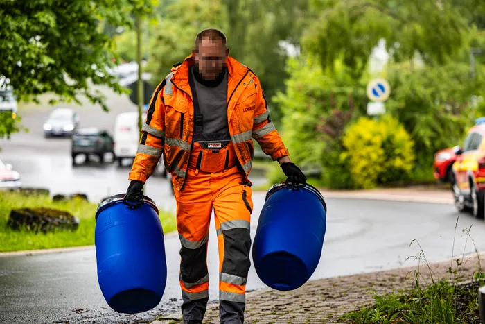 Einsatzkraft in orangefarbener Schutzkleidung trägt zwei blaue Fässer an einer Straße mit Einsatzfahrzeugen im Hintergrund. Im Hintergrund sind Bäume und Büsche zu sehen.