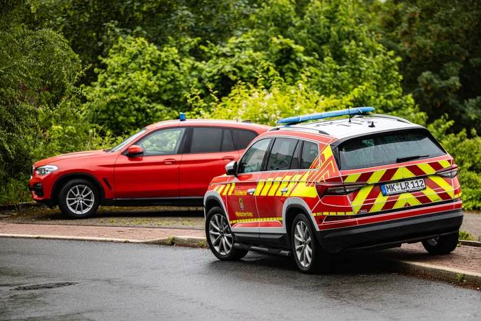 Zwei rote Einsatzfahrzeuge der Feuerwehr stehen auf einer nassen Straße vor Grünflächen. Im Hintergrund ist ein rotes Auto geparkt.