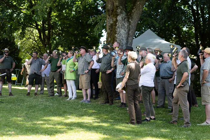 Eine Gruppe von etwa 30 Personen, hauptsächlich Männer mittleren Alters, steht auf einer grünen Wiese unter einem großen Baum und spielt auf Blechhörnern. Einige Personen schauen dem Dirigenten zu, der mit dem Rücken zum Betrachter steht. Im Hintergrund ist ein Zelt zu sehen.