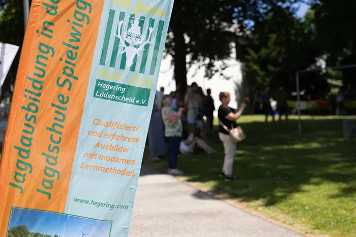 Ein vertikales Banner mit grün-weißem Streifenmuster und einem Hirschmotiv kündigt die Jagdschule Spielwig an. Darunter stehen Informationen zum 100-jährigen Bestehen des Hegerings Lüdenscheid und zur Ausbildung. Im Hintergrund sind verschwommene Personen auf einer grünen Wiese zu sehen.