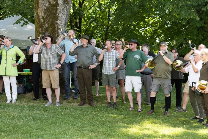 Eine Gruppe von etwa 20 Personen spielt auf Blechhörnern und Posaunen im Freien auf einer grünen Wiese. Einige tragen traditionelle Kleidung, darunter eine Frau in einem grünen Dirndl.