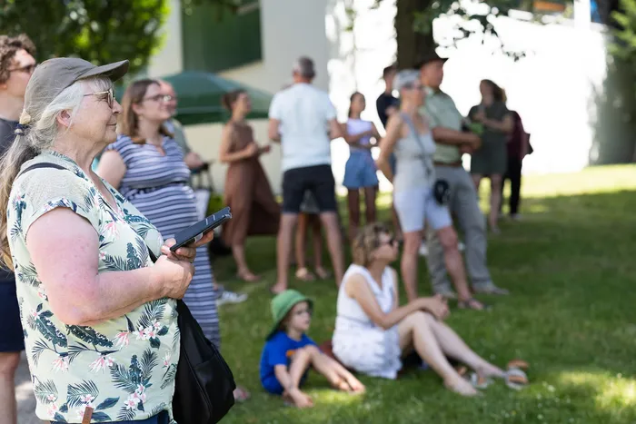 Eine Frau mit grauen Haaren und einer Baseballkappe steht im Vordergrund und hält ein Notizbuch und einen Stift. Im Hintergrund sitzen und stehen Menschen auf einer grünen Wiese, vermutlich bei einer Feier.