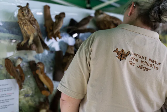 Eine Person mit einem beigefarbenen T-Shirt, auf dem „Lernort Natur – eine Initiative der Jäger“ steht, steht vor geschnitzten Holzfiguren von Vögeln und anderen Tieren. Im Hintergrund sind weitere Holzskulpturen und ein Zelt zu sehen.