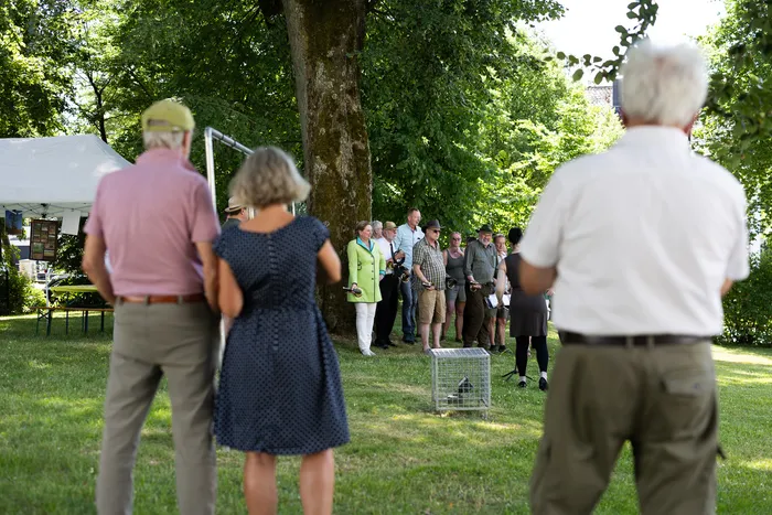 Eine Gruppe von Menschen steht auf einer Wiese vor einem großen Baum. Einige tragen traditionelle Jagdkleidung mit grünen Jacken und Hüten. Im Hintergrund ist ein Festzelt zu sehen.