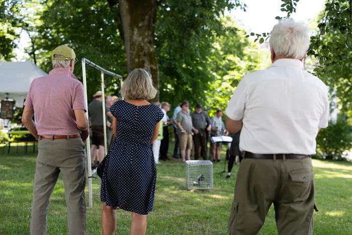 Zuschauer stehen dem Rücken zugewandt und beobachten eine Gruppe von Personen, die vor einem Baum auf einer Wiese stehen. Im Hintergrund ein Zelt und Bäume, die Szene zeigt eine Feier zum 100-jährigen Bestehen des Hegerings.