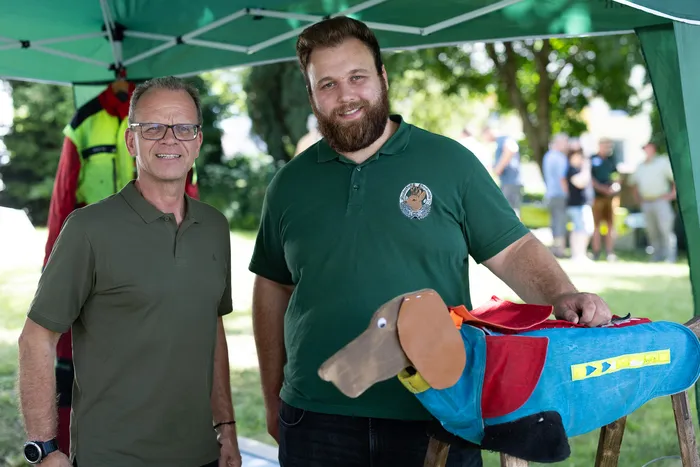 Zwei Männer lächeln in die Kamera. Der Mann links trägt ein rotes Poloshirt und eine Smartwatch, der Mann rechts ein grünes Poloshirt mit Logo und hält ein Stofftier in Form eines Hundes. Im Hintergrund ein Zelt und weitere Personen.
