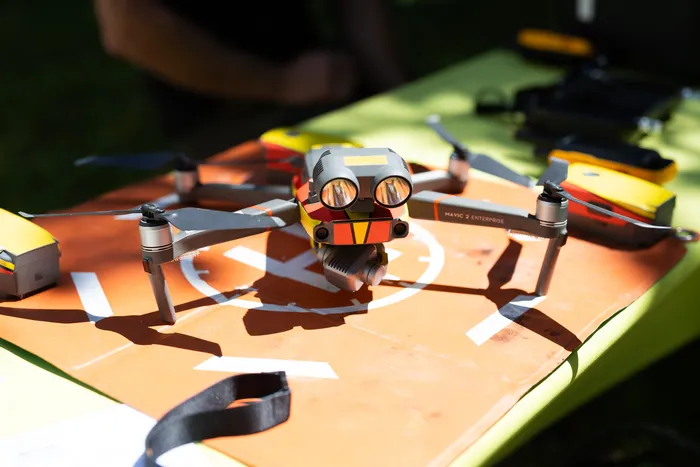 Two drones with attached faces and sunglasses are positioned on an orange landing pad with a target design. A black strap lies in the foreground, and blurred figures and objects are visible in the background.
