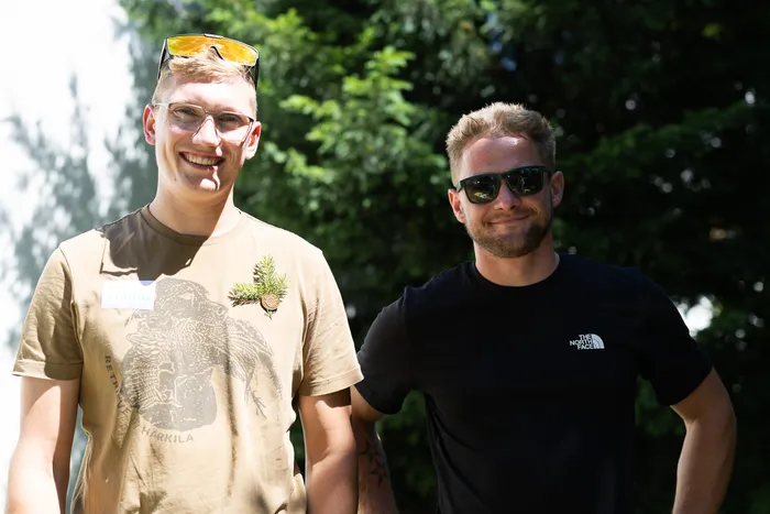Zwei lächelnde Männer stehen im Freien vor grüner Vegetation. Der Mann links trägt eine braune T-Shirt mit einem aufgedruckten Adler, der rechte eine schwarze T-Shirt mit Logo.