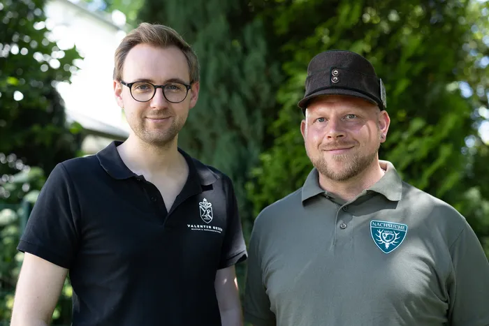 Zwei Männer im Poloshirt stehen lächelnd vor grüner Vegetation. Der Mann links trägt eine Brille und ein schwarzes Poloshirt mit Logo, der Mann rechts eine Kappe und ein olivgrünes Poloshirt mit aufgesticktem Wappen.