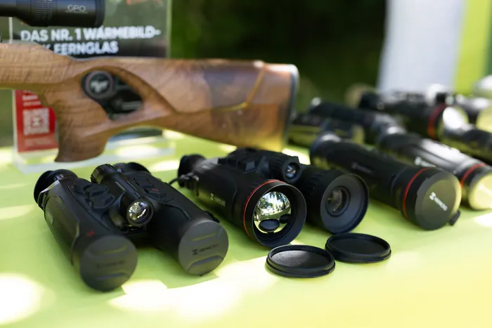 Mehrere Wärmebildfernrohre und ein Gewehr liegen auf einem Tisch mit hellgrünem Tuch. Im Hintergrund ist ein Banner mit der Aufschrift „DAS NR. 1 WÄRMEBILD-FERNGLAS“ zu sehen.