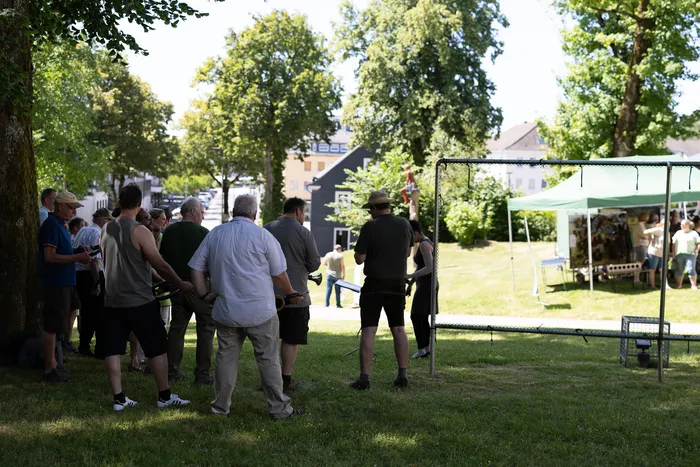 Eine Gruppe von Männern steht auf einer Wiese vor einem Volleyballnetz und beobachtet etwas. Im Hintergrund sind weitere Personen und ein Festzelt zu sehen.