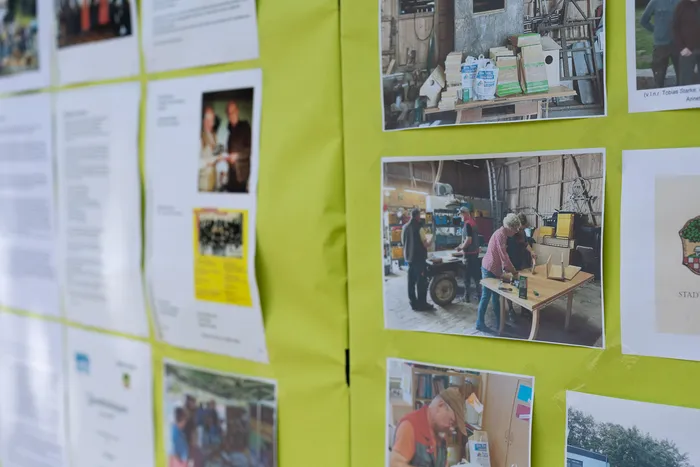 Eine Pinnwand mit Fotos von Menschen und Aktivitäten im Freien, darunter eine Frau, die an einem Tisch arbeitet, und eine Gruppe, die zusammensteht. Die Fotos scheinen verschiedene Aspekte eines hundertjährigen Jubiläums zu zeigen.