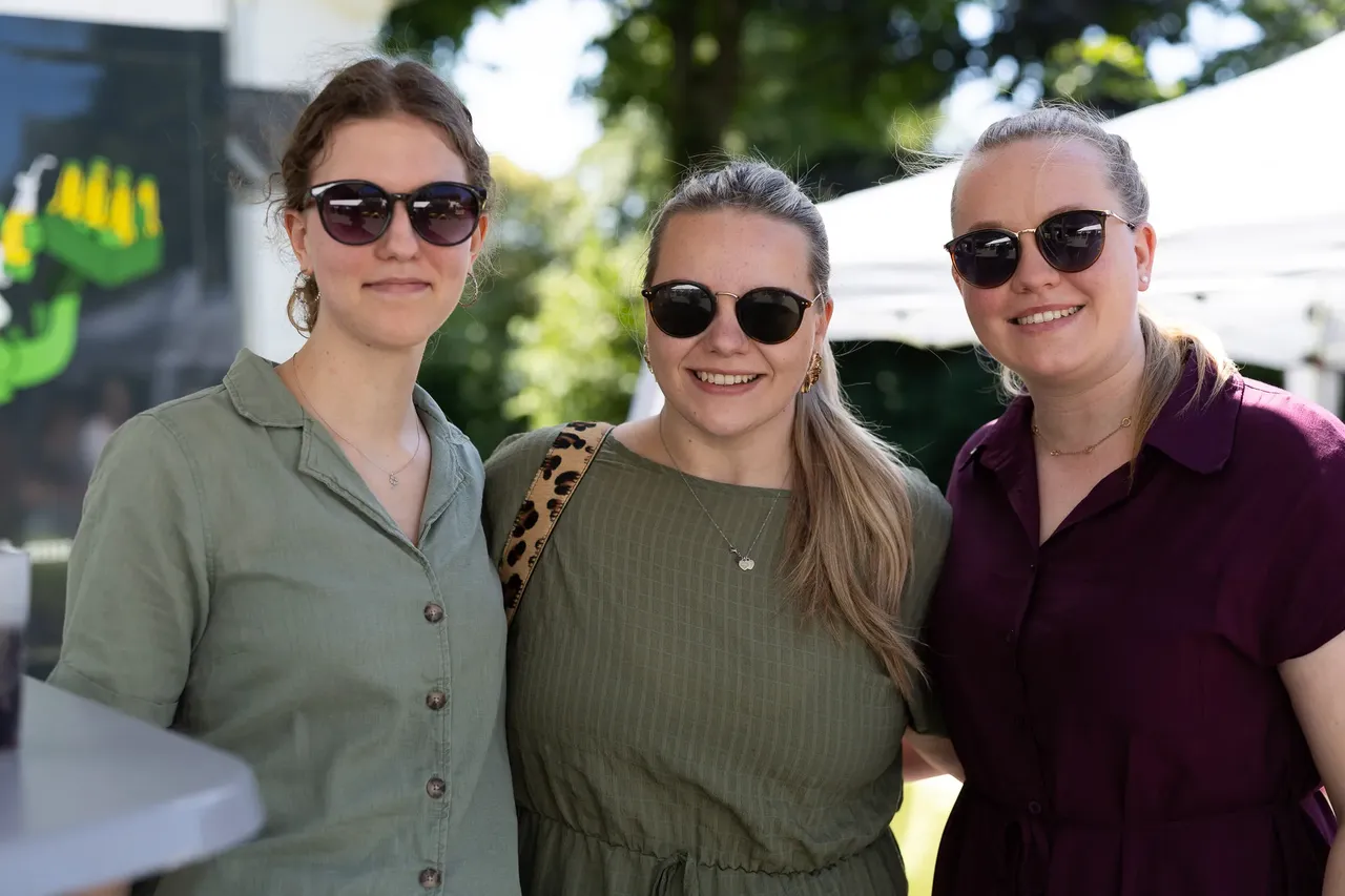 Drei lächelnde Frauen mit Sonnenbrillen stehen eng nebeneinander. Sie tragen sommerliche Kleidung und Schmuck. Im Hintergrund ist grünes Laub zu sehen.