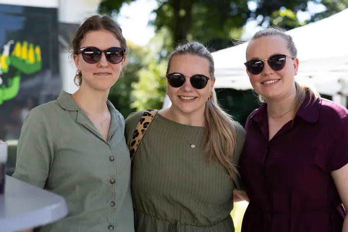 Drei lächelnde Frauen mit Sonnenbrillen stehen eng nebeneinander. Sie tragen sommerliche Kleidung und Schmuck. Im Hintergrund ist grünes Laub zu sehen.