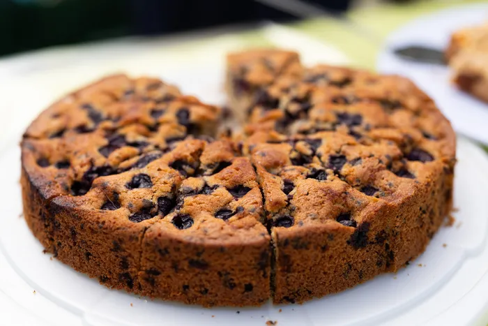 Ein Stück Blaubeerkuchen auf einem weißen Teller. Der Kuchen ist bereits angeschnitten und zeigt viele dunkle Blaubeeren im Teig.