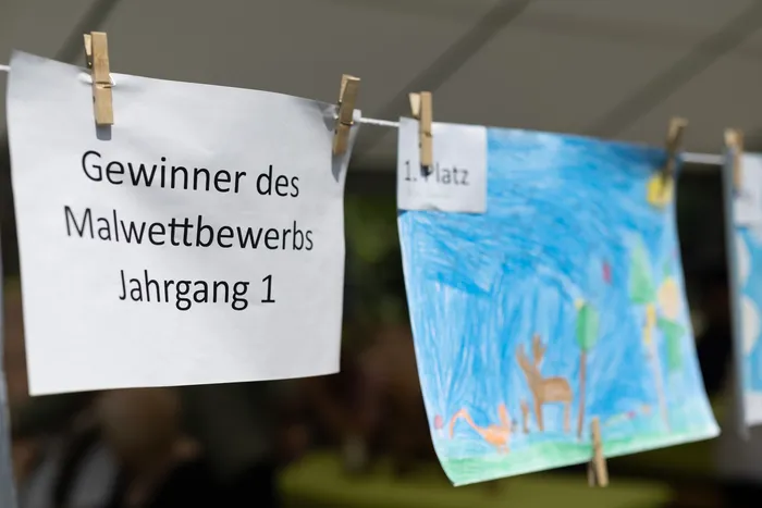 Gewinnerbild des Malwettbewerbs Jahrgang 1 hängt mit einer Wäscheklammer an einer Leine, daneben ein weiteres Bild mit der Aufschrift '1. Platz'. Die Bilder zeigen Kinderzeichnungen.
