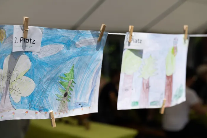 Kinderzeichnungen, befestigt an einer Wäscheleine mit Wäscheklammern, zeigen farbenfrohe Motive. Ein Schild kennzeichnet die Bilder als '2. Platz' und '3. Platz'.