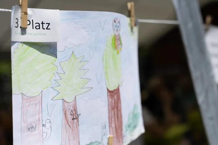 Ein Kinderbild hängt an einer Wäscheleine, beschriftet mit „3. Platz“ und dem Namen „Aue Horschkora“. Das Bild zeigt Bäume und Tiere in leuchtenden Farben.