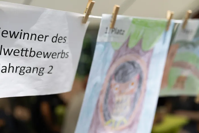 Kinderzeichnungen, befestigt mit Wäscheklammern an einer Leine, zeigen verschiedene Motive und sind mit Beschriftungen wie „Gewinner des Wettbewerbs“ und „1. Platz“ versehen.